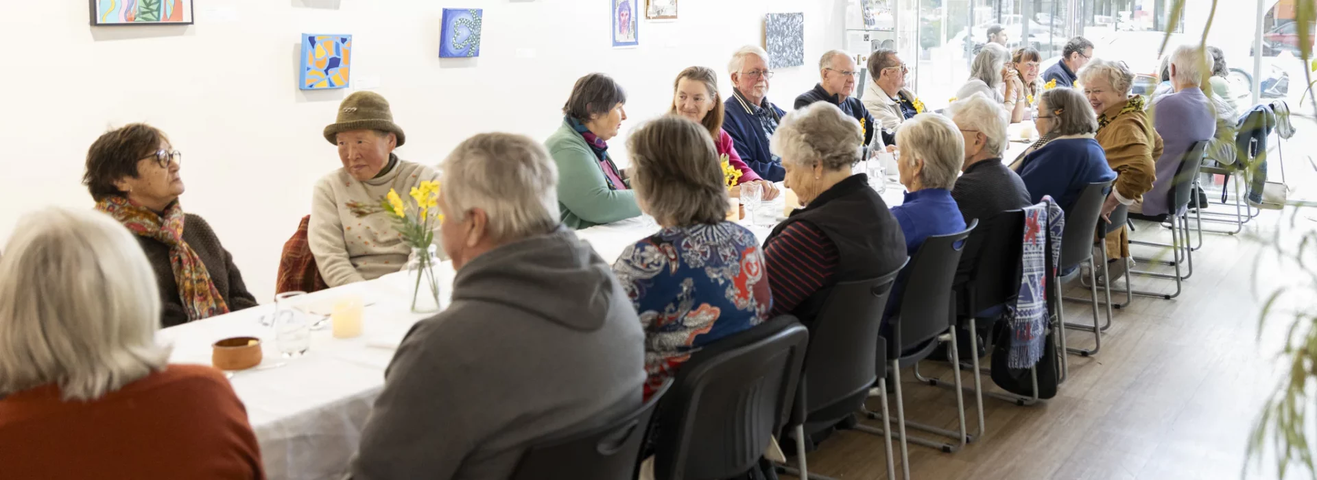 A group of people sits at long tables in a brightly lit room, engaging in conversation. Artwork decorates the white walls, and small vases with flowers adorn the tables. The atmosphere is casual and social.