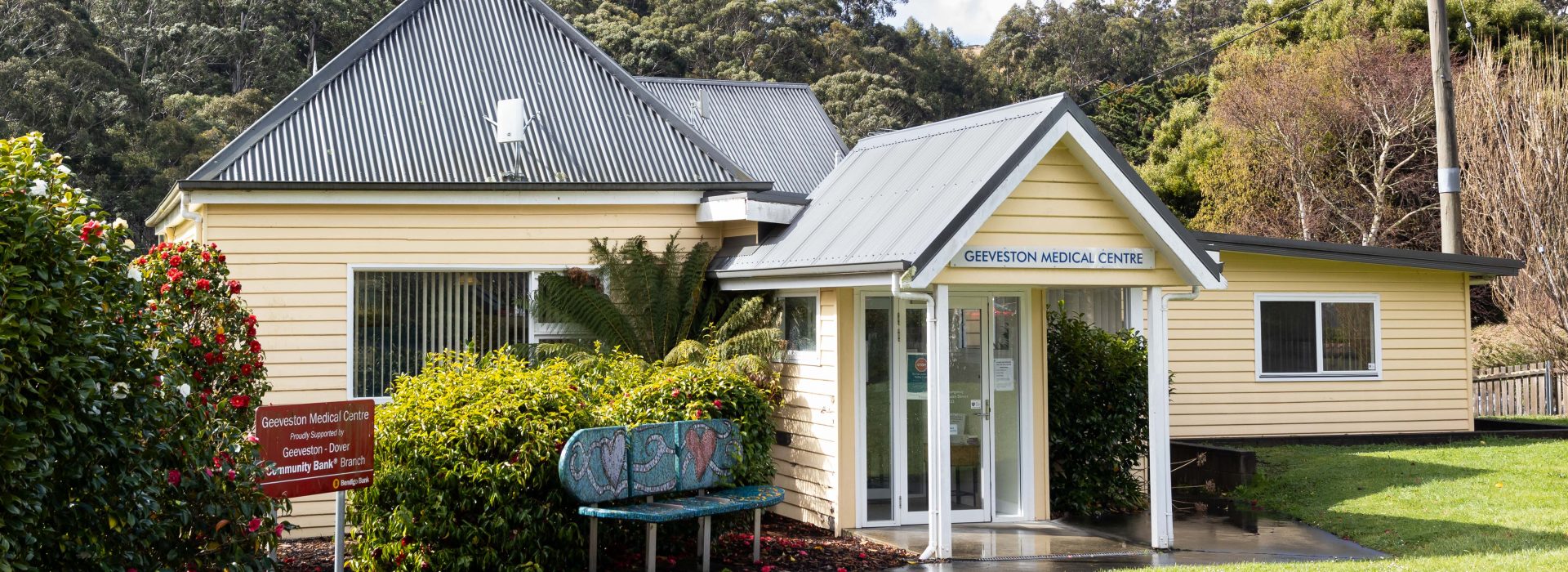 A small, yellow medical center with a metal roof is surrounded by greenery. A sign reads Geeveston Medical Centre. Theres a bench outside, and trees fill the background under a cloudy sky. A paved path leads to the entrance.