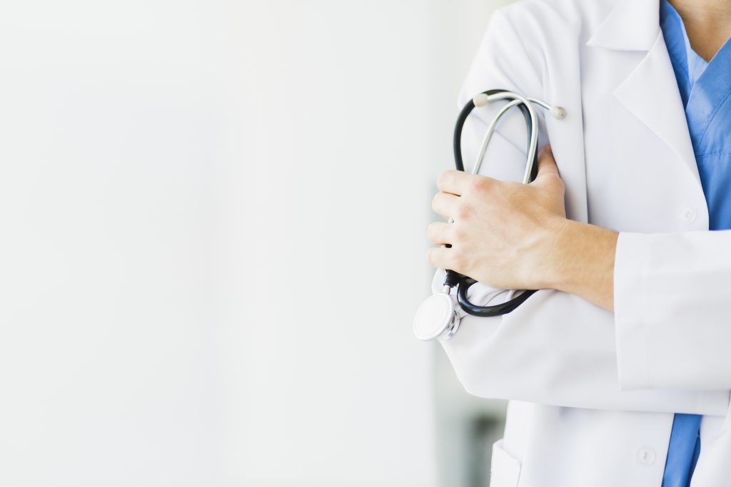 A healthcare professional in a white coat stands with arms crossed, holding a stethoscope. The background is softly blurred, emphasizing the focus on the doctor and their attire. The image conveys a sense of medical expertise.