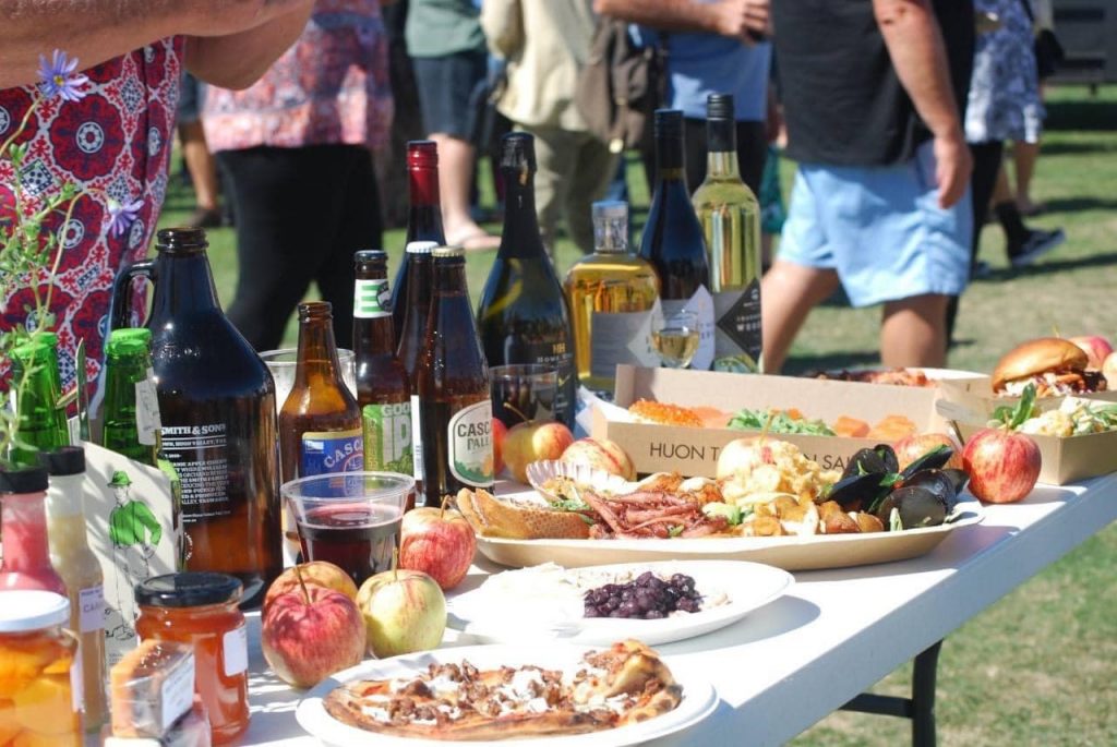 A table outdoors is filled with various foods and drinks, including bottles of wine, beer, apples, plates of pizza, and appetizers. People stand in the background, slightly out of focus, enjoying a sunny day.