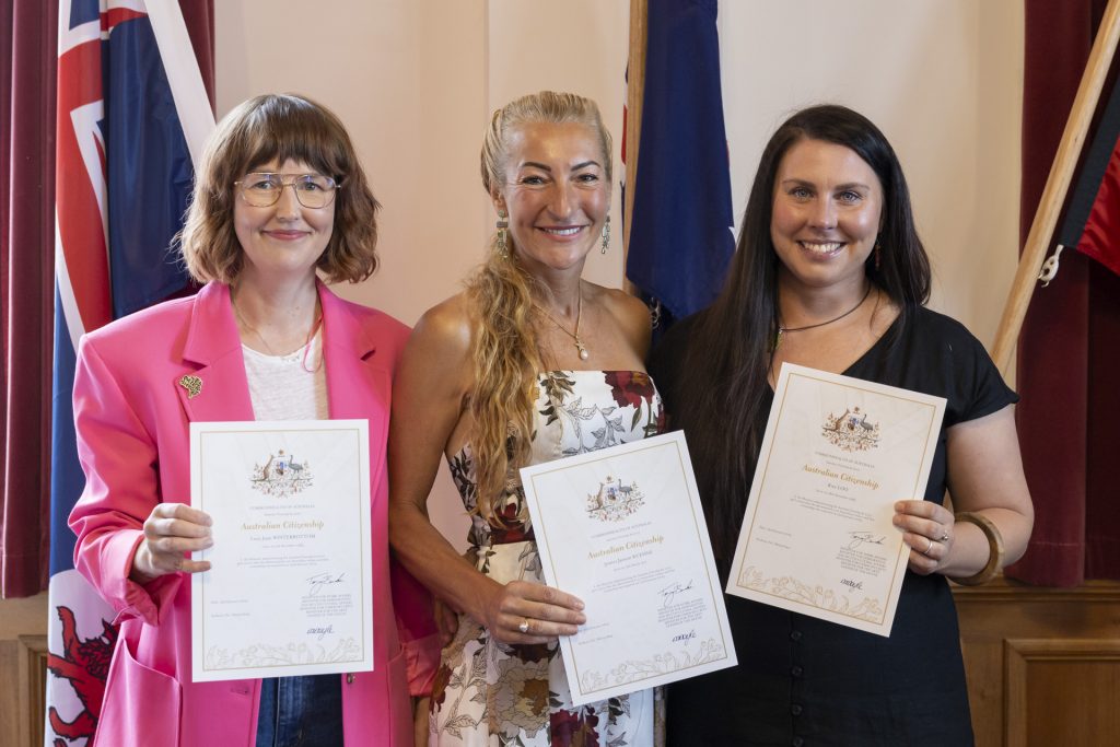 Three people smile while holding certificates. The person on the left wears a pink blazer, the person in the middle wears a floral dress, and the person on the right wears a black top. Flags are visible in the background.