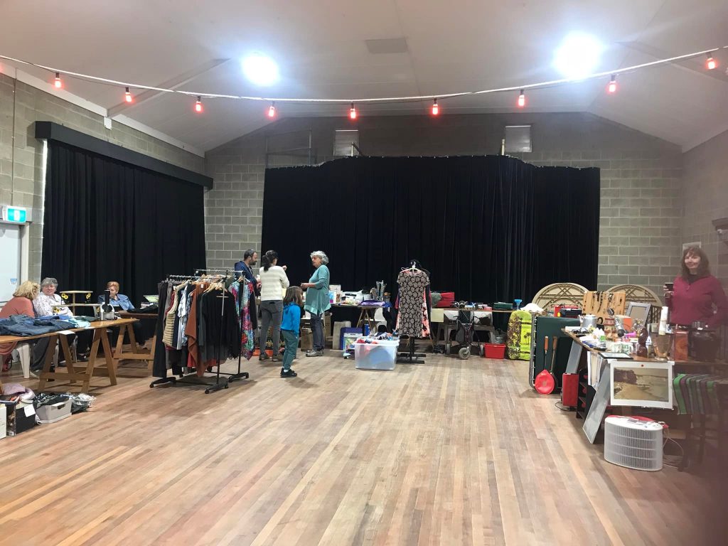 A community hall with wooden floors, where people are browsing items like clothes and crafts displayed on tables and racks. Red string lights hang across the ceiling, and various stalls are set up along the walls with people attending each.