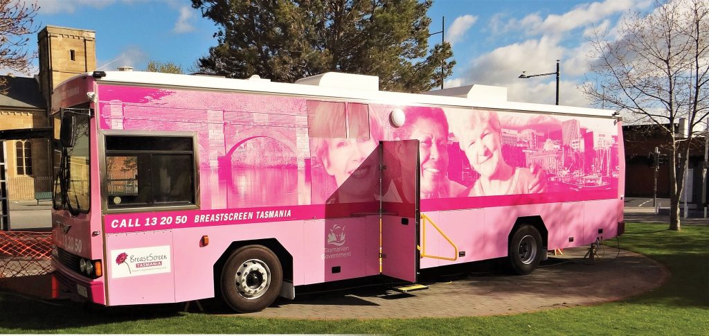 A pink mobile screening bus with large images of women is parked on grass. The text BreastScreen Tasmania and Call 13 20 50 are visible. The bus is named Luna. Trees and a building are in the background under a blue sky.