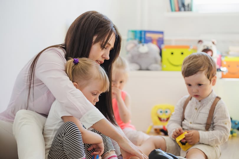 A woman and three young children are sitting on the floor in a playroom. The woman is interacting with a girl in her lap, while a boy in suspenders plays nearby. Toys and colorful decor are visible in the background.