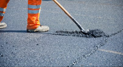 A worker in orange reflective pants uses a shovel to spread asphalt on a road. The focus is on the lower half of the workers body and the shovel. The asphalt appears fresh and black against the road surface.