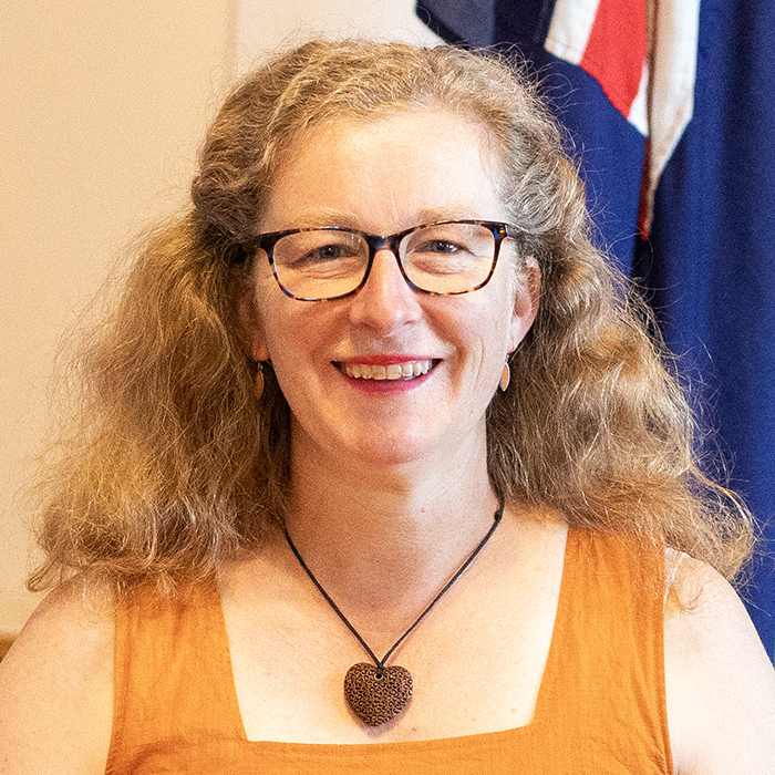 Woman with curly hair, wearing glasses and a brown sleeveless top, smiles at the camera. She has a heart-shaped necklace. A flag is visible in the background.