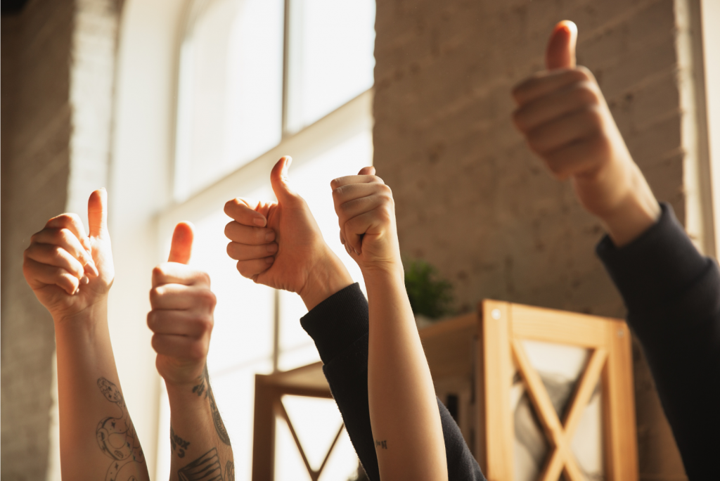 Five hands giving a thumbs-up gesture, raised in the air. The hands vary in skin tone and have tattoos. Soft lighting from nearby windows illuminates the scene, with a blurred background featuring a brick wall and wooden shelves.