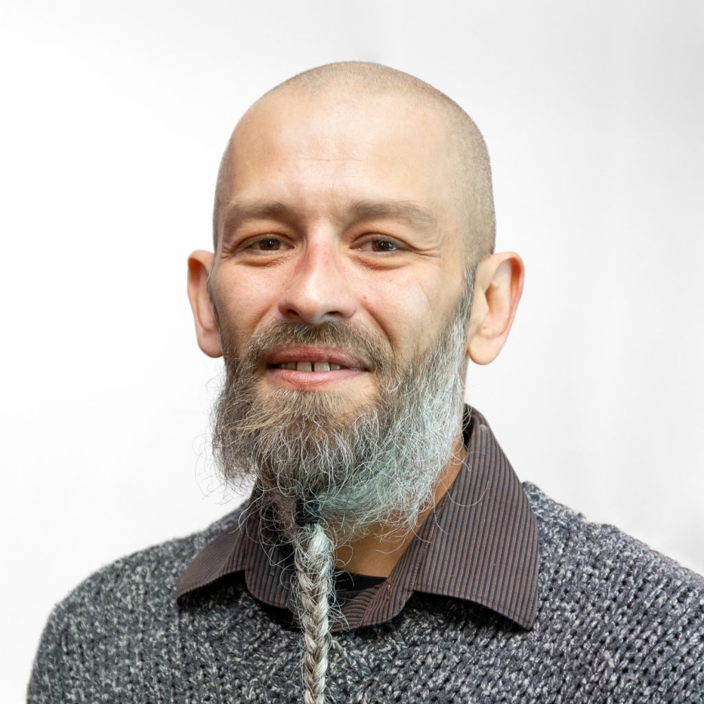 A person with a beard and braided strand smiles, wearing a gray sweater over a collared shirt. The background is plain white.