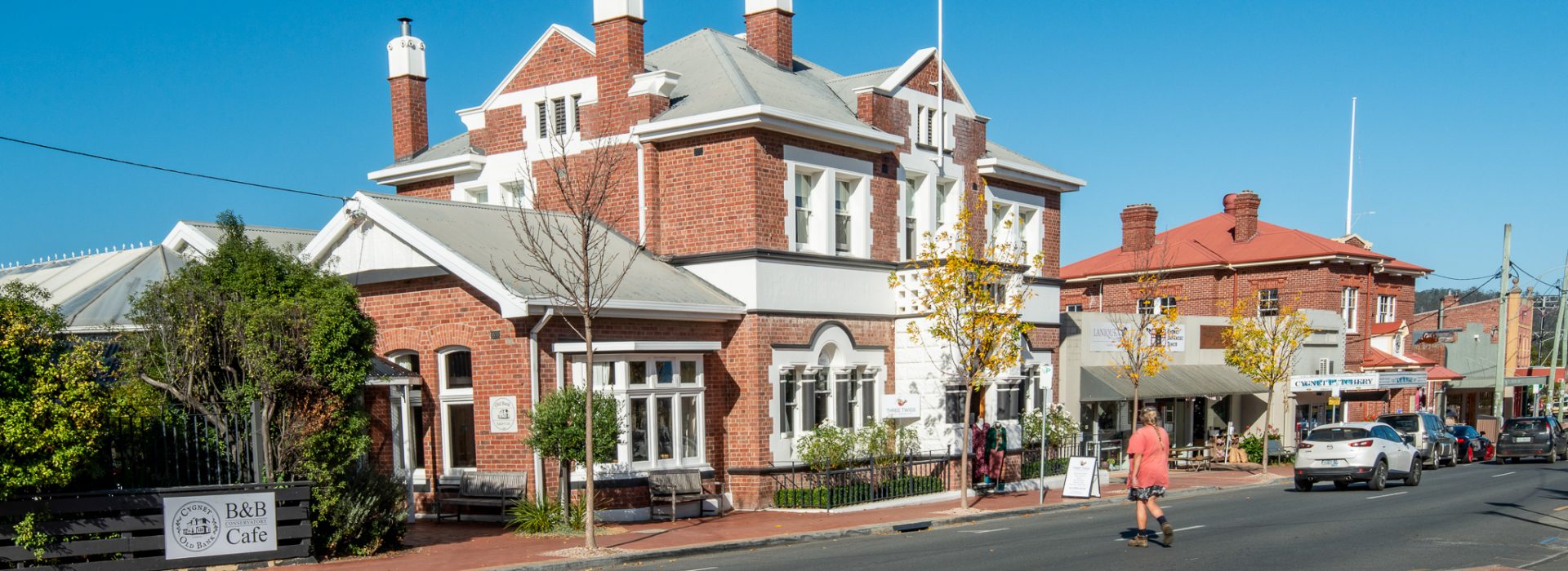 Street scene with a red-brick building featuring chimneys and a flag on the corner. A B&B cafe sign is visible. A person is walking on the sidewalk, and there are parked cars and other buildings in the background under a clear blue sky.