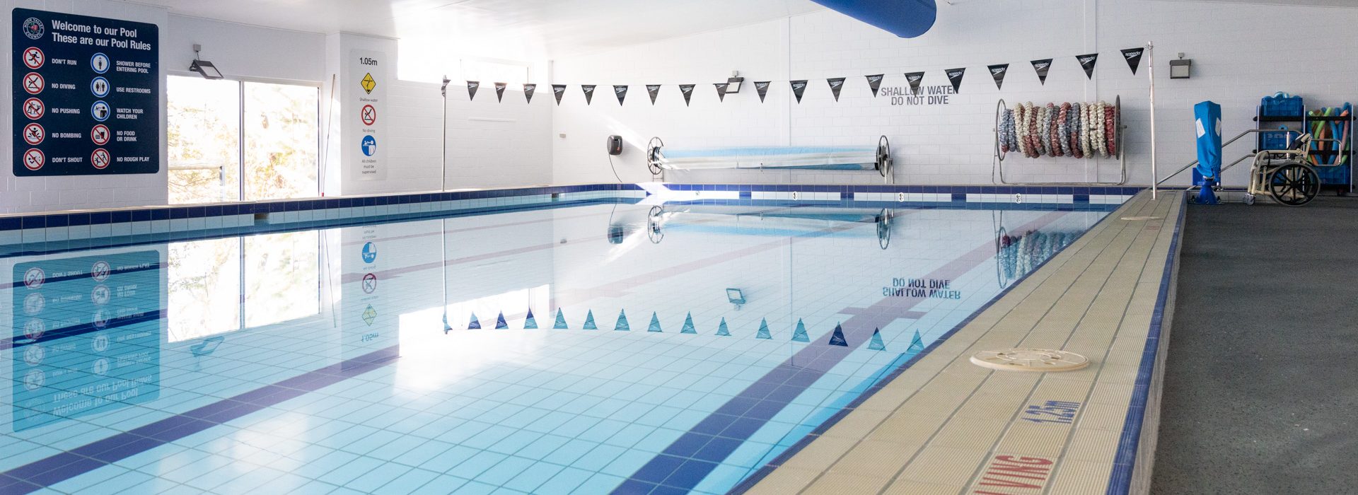 Indoor swimming pool with clear water and tiled floor. Safety signs and swimming lane dividers are visible. A large blue ventilation duct runs along the ceiling, and a stack of pool noodles is on the far wall next to a wheelchair-accessible lift.