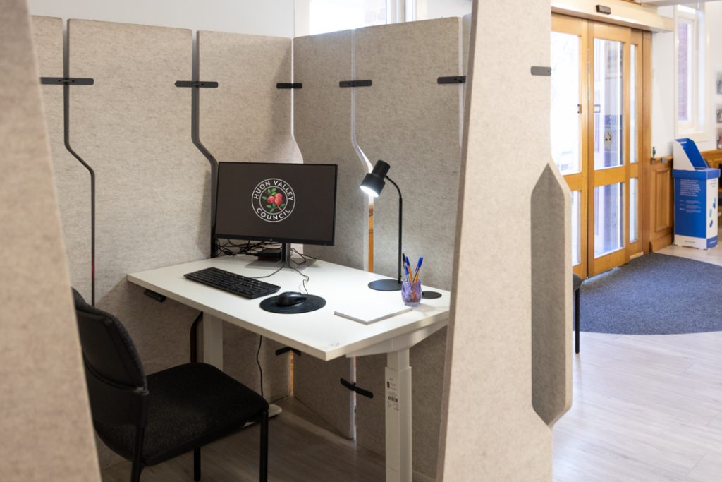 A small study booth with a computer, keyboard, and mouse on a white desk. A lamp is next to the computer, along with pens in a holder. The booth is partitioned with gray panels. A bright doorway is visible in the background.