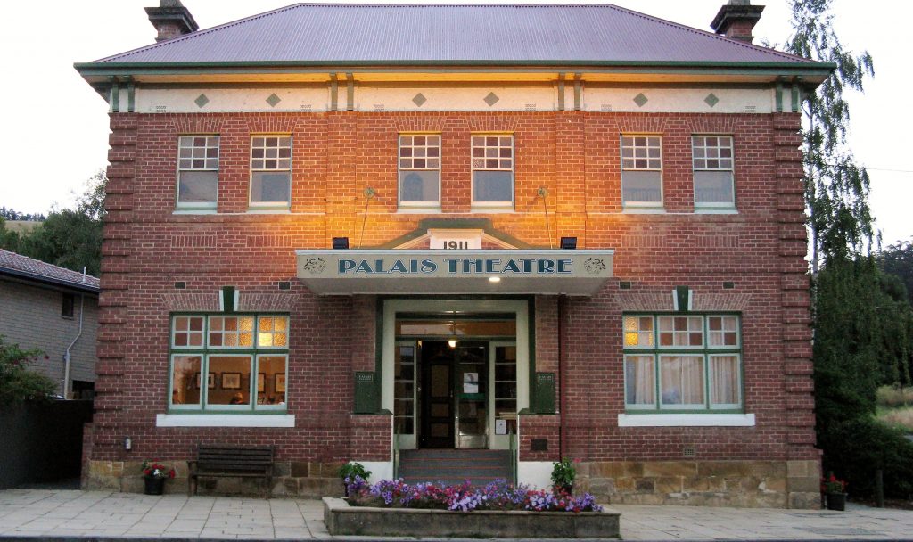 A historic brick building labeled Palais Theatre with an entrance lit by warm lights. It features large windows, a small garden with flowers, and a bench in front. The buildings architecture is traditional with a visible date 1911 above the entrance.