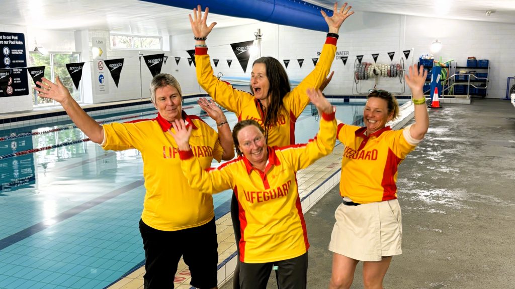 Four lifeguards in yellow and red uniforms pose joyfully by an indoor swimming pool. They are standing on the pool deck, raising their arms and smiling widely. The pool and flags are visible in the background.