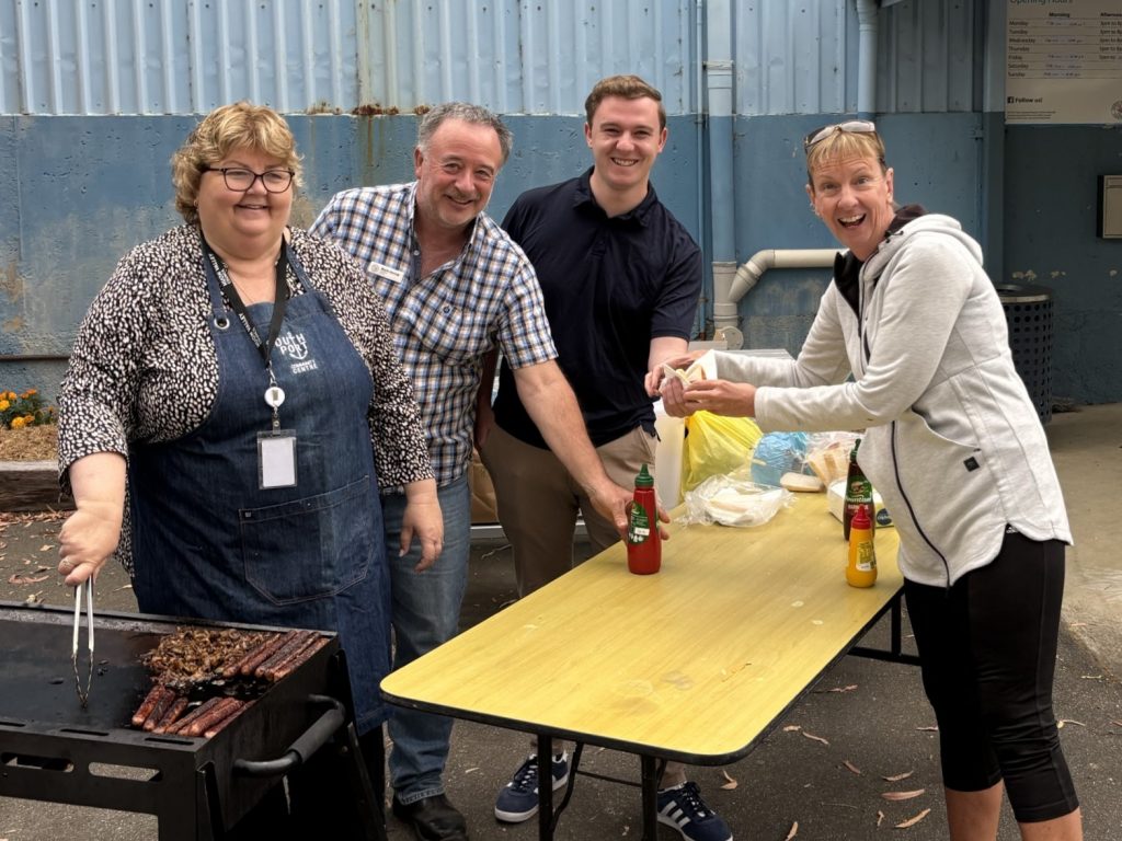 Four people gather around a grill and table outdoors, smiling as they prepare food. On the table are condiments and bags. The setting appears casual and friendly, with a blue corrugated wall in the background.