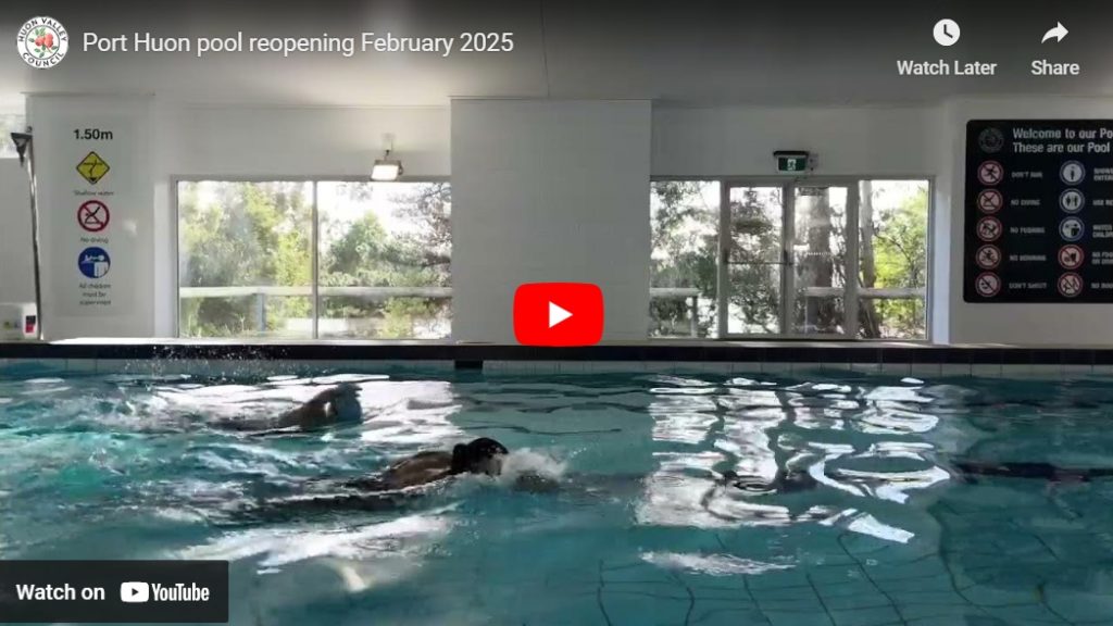 Two people swim in an indoor pool under bright lighting. Large windows show trees outside. Safety signs and a pool depth marker are visible. A video title at the top reads Port Huon pool reopening February 2025. A YouTube play button is in the center.