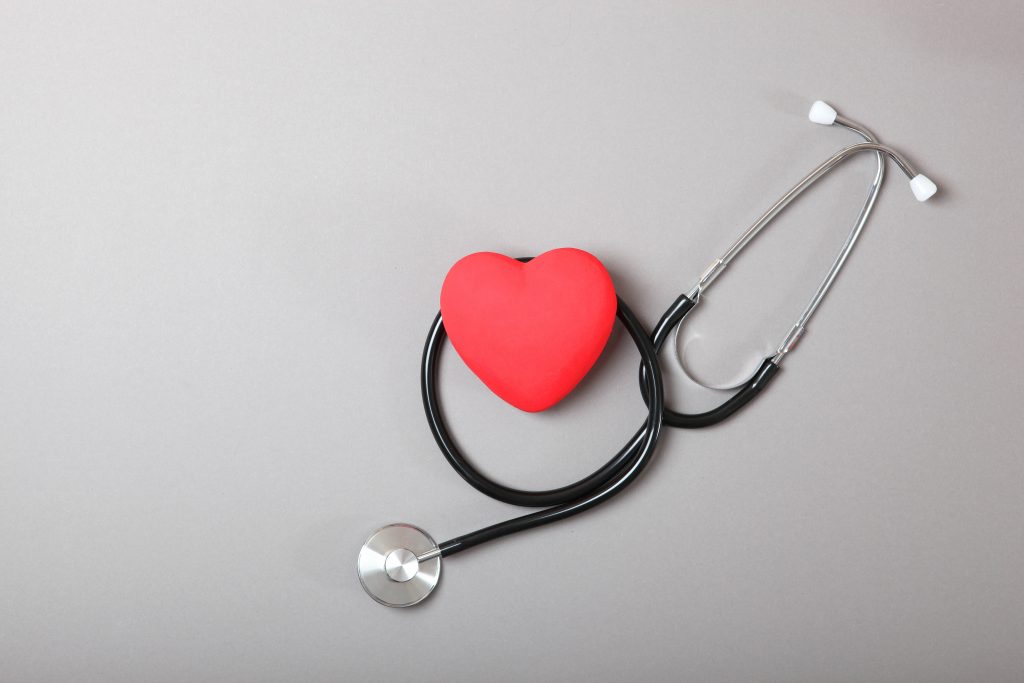 A red heart-shaped model is placed above a stethoscope on a gray background, symbolizing healthcare and cardiology. The stethoscope is partially coiled around the heart.