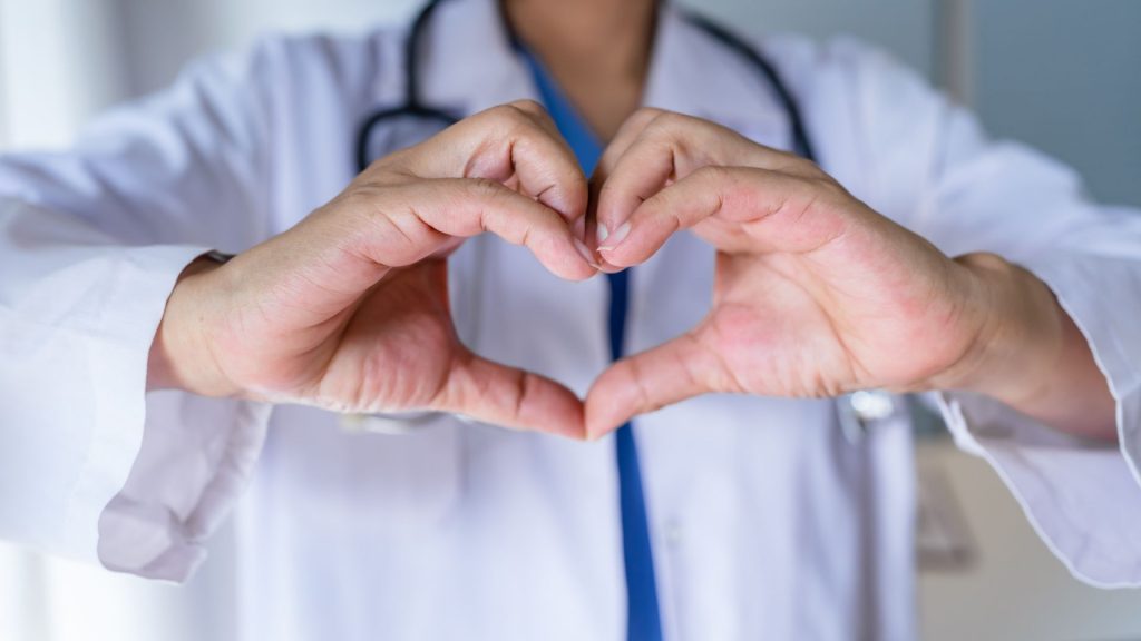 A doctor wearing a white coat and stethoscope forms a heart shape with their hands. The focus is on the hands in the heart shape, with the face out of frame. The background is blurred.