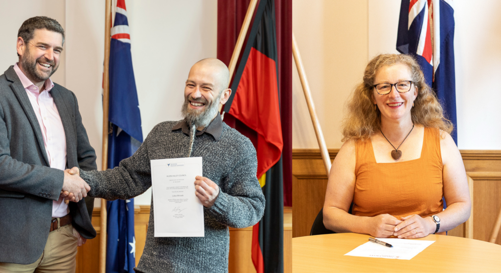 A man in a suit shakes hands with a smiling man holding a certificate in front of Australian and Indigenous flags. A woman with glasses and curly hair sits at a table, smiling, with a document in front of her. Flags are visible in the background.