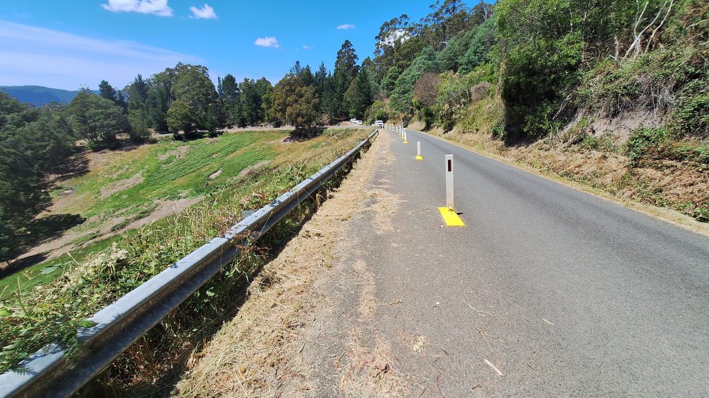 A winding road stretches through a lush, hilly landscape. Trees line both sides, and a guardrail runs alongside the pavement. The sky is clear with a few clouds, and the road has markers placed in the center for safety.