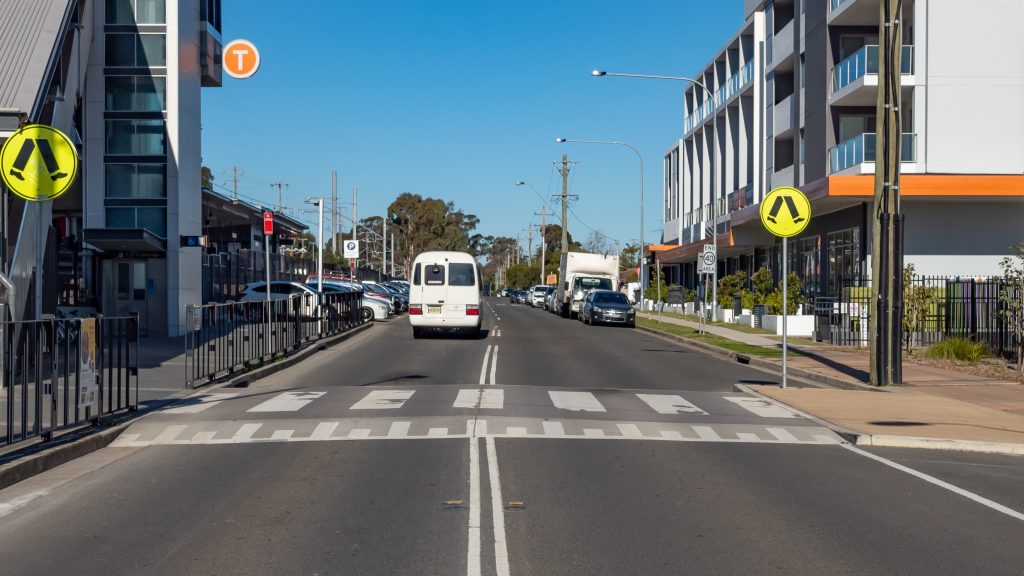 A white van drives down a city street flanked by buildings and parked cars. Pedestrian crossing signs are visible on both sides of the road. A clear blue sky provides a backdrop, with a large orange T sign on the left.