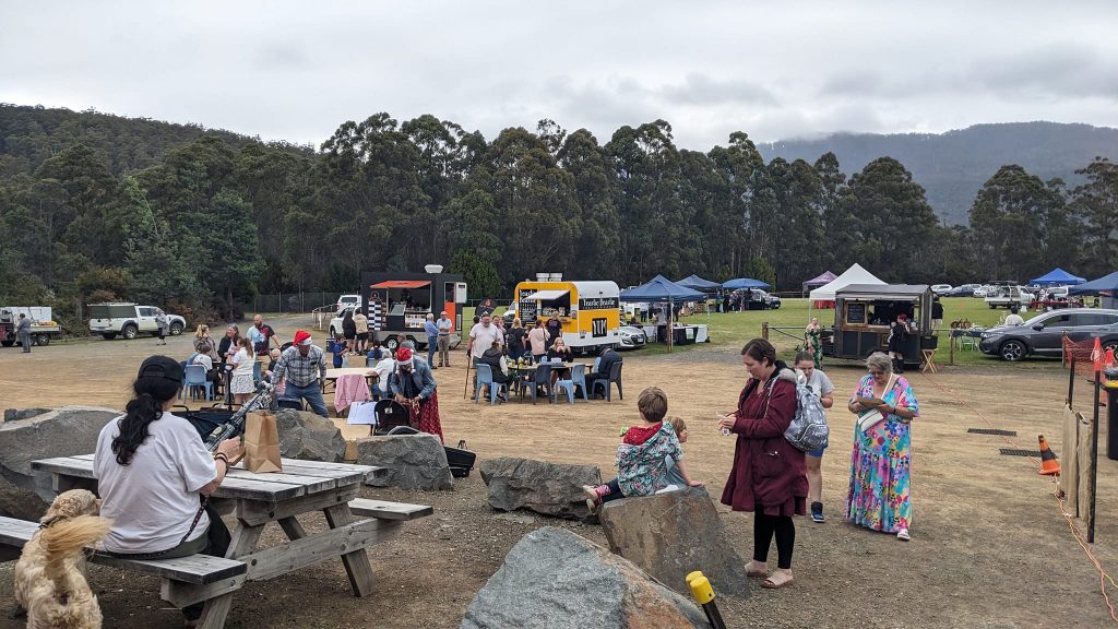 People gather at an outdoor market with food trucks, tents, and picnic tables. A cloudy sky and trees form the backdrop. Attendees, including families, engage in conversations and explore the area.