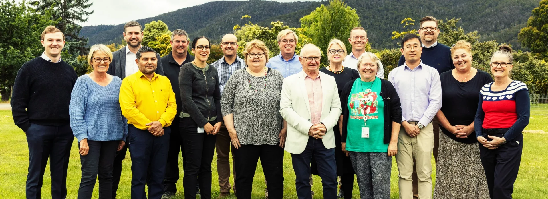 A group of people standing in a grassy area with trees and mountains in the background. Most are smiling. They are wearing a variety of clothing styles, including shirts, sweaters, and a Christmas-themed t-shirt. Its a cloudy day.