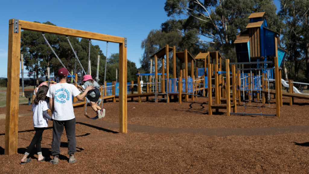 A man pushes a child on a swing at a playground, with another child standing close by. The playground features multiple climbing structures and slides, surrounded by wood chips and trees under a clear blue sky.