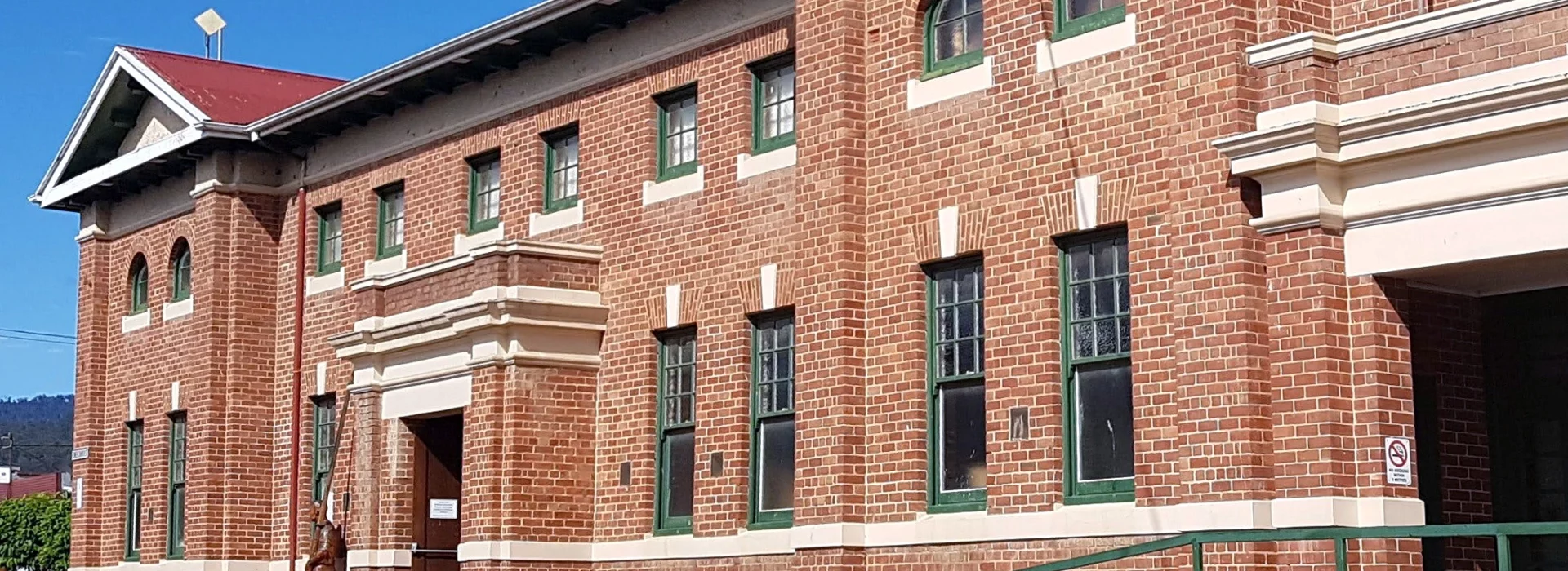 A large brick building featuring arched windows and decorative columns. The structure has a maroon roof and a green access ramp. The sky is clear and blue.