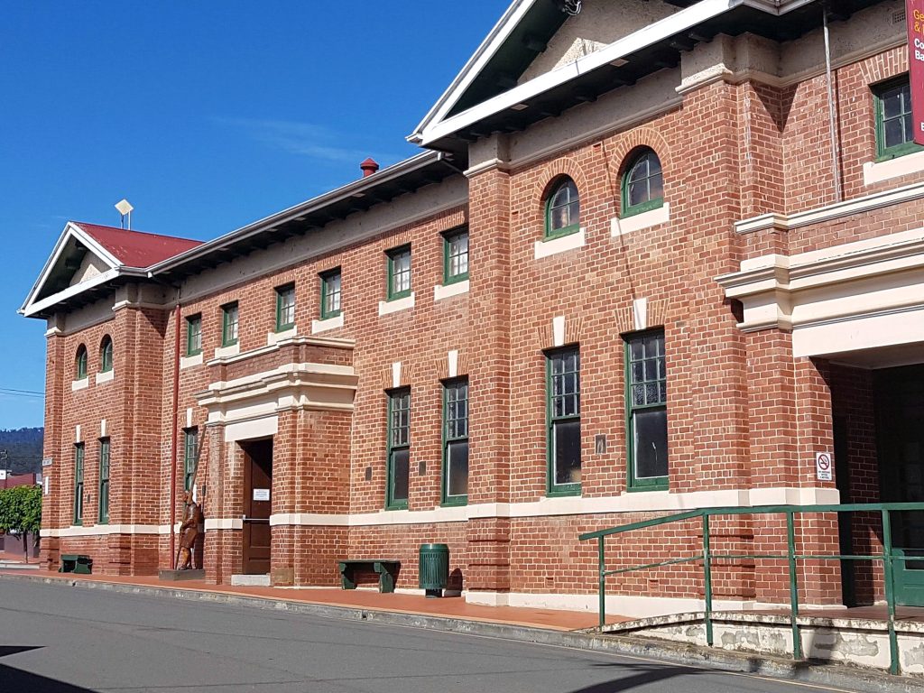 A brick building with green and white trim, featuring multiple windows and arched details. There is a red roof and a white decorative cornice. A green ramp and steps lead to the entrance. Clear blue sky in the background.