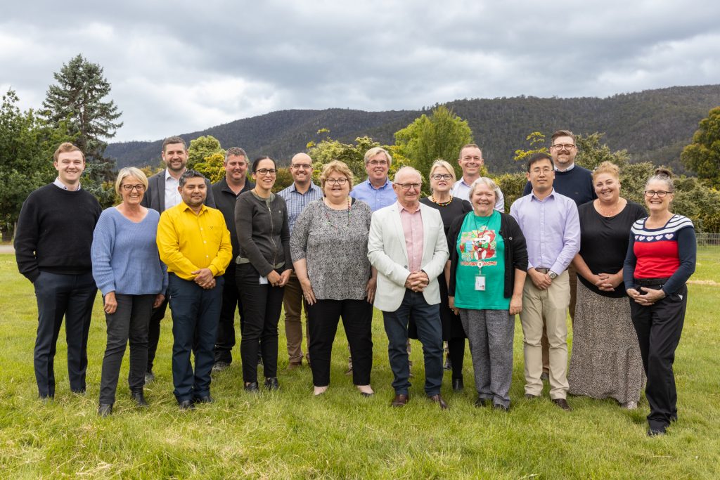 A group of fifteen people standing outdoors on grass with trees and hills in the background. They are dressed in casual and business attire and smiling at the camera. The sky is cloudy.