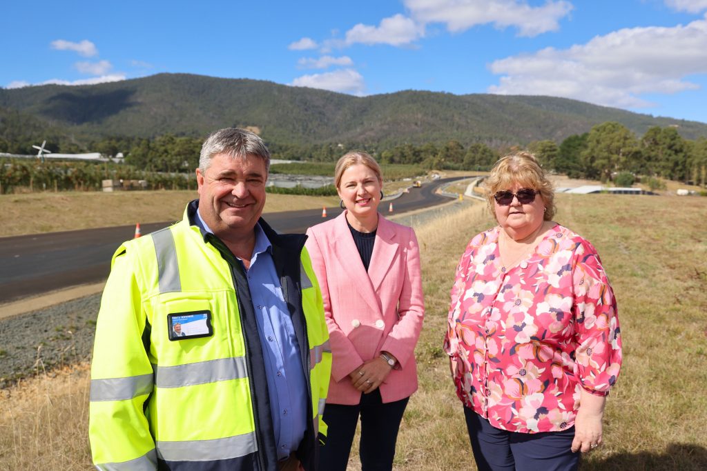 Three individuals stand outdoors on a sunny day. The first, wearing a high-visibility jacket, smiles alongside a woman in a pink blazer and another in a floral blouse. A newly paved road and forested hills are in the background under a blue sky.