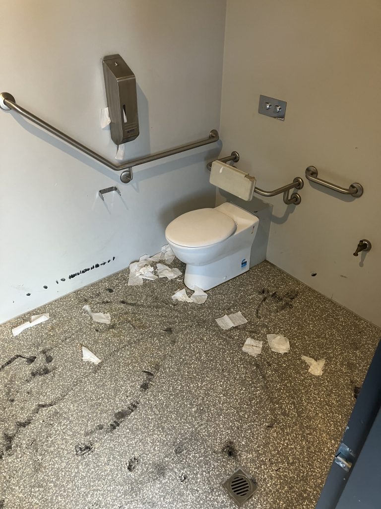 A public restroom with a toilet and grab bars around it. The floor is strewn with crumpled paper towels and has footprints. A toilet paper holder is mounted on the wall, and the scene is generally untidy.