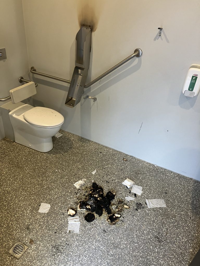 A public restroom with a burned section on the wall around a damaged dispenser. Charred debris and scattered tissues lie on the floor, indicating a fire incident. The bathroom has a toilet, handrails, and a wall-mounted sanitizer.